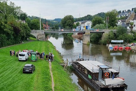 Ein großes Floß liegt vertäut am Ufer der Lahn. Daneben warten ein Transporter und ein PKW und einige Menschen.