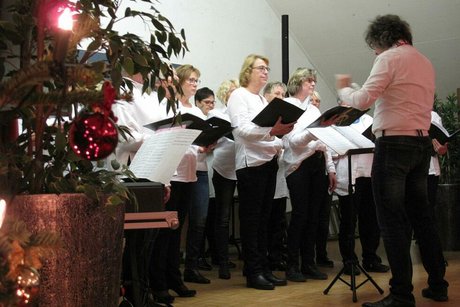 Der Chorleiter dirigiert singende Frauen, die auf einer Bühne stehen. Links im Bild befindet sich ein Weihnachtsbaum.