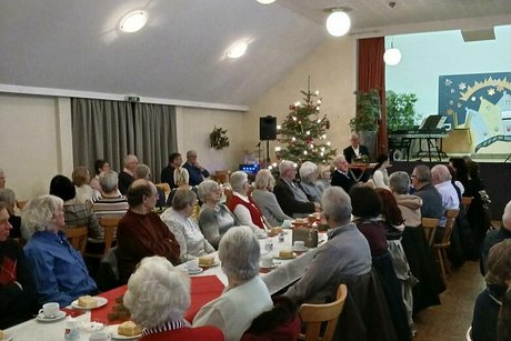 Viele Menschen sitzen in einem Saal an langen, weihnachtlich geschmückten Tischen.