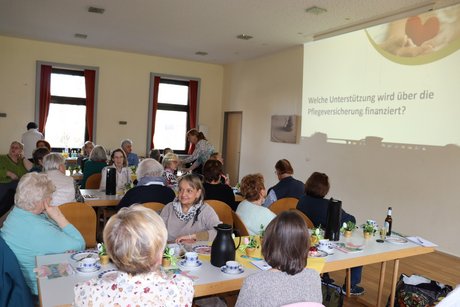 Treffen der Vertreterinnen der Frauen im Mehrgenerationenhaus. Mehrere Menschen an langen Tischen verfolgen den Vortrag und die Präsentation zum Thema Pflegeversicherung.