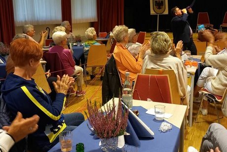 Klatschende Menschen sitzen an Tischen. Im Hintergrund zeigt ein Zauberer Ballonkunst.