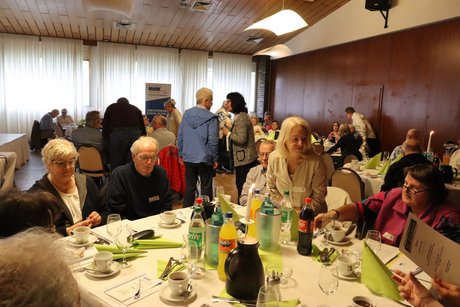 Im Vordergrund sitzen mehrere Personen an einem Tisch. Im Hintergrund sieht man Menschen an anderen Tischen sitzen und sich auch stehend zu unterhalten