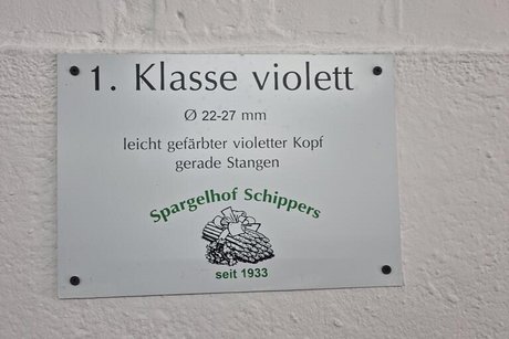 1. Klasse violett steht auf einem Metallschild, das beim Spargelhof an einer weißen Wand angebracht ist.