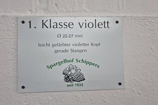 1. Klasse violett steht auf einem Metallschild, das beim Spargelhof an einer weißen Wand angebracht ist.