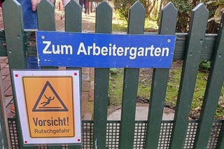 Ein Schild mit der Aufschrift Zum Arbeitergarten hängt an einem grünlackierten Lattenzaun. Darunter weist ein weiteres, gelbes Warnschild mit Piktogramm eines fallenden Menschen auf Rutschgefahren hin.