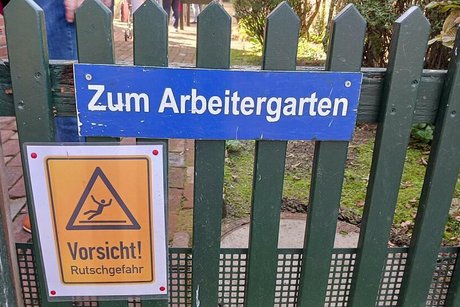 Ein Schild mit der Aufschrift Zum Arbeitergarten hängt an einem grünlackierten Lattenzaun. Darunter weist ein weiteres, gelbes Warnschild mit Piktogramm eines fallenden Menschen auf Rutschgefahren hin.