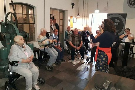 Eine Gruppe Menschen steht oder sitzt auf Rollatoren um alte Maschinen herum und hört einer Frau zu, die etwas erzählt.