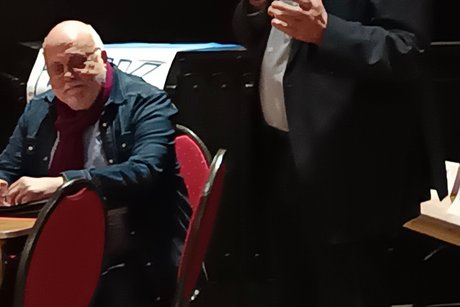 Auf dem Bild sieht man einen Mann im dunklen Anzug mit Mikrofon in der Hand der gerade zu den Mitgliedern spricht.