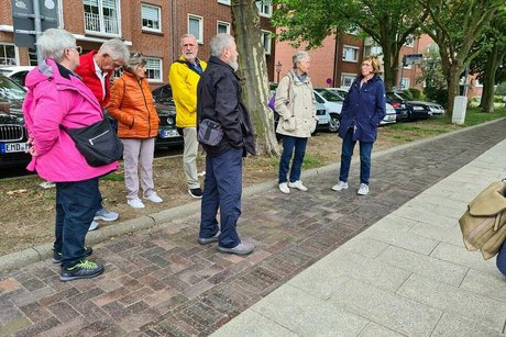 Menschen in bunten Regen- und Outdoorjacken stehen auf einem von Bäumen gesäumten Gehweg.