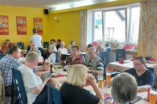 Menschen sitzen gemeinsam an Tischen im Restaurant der Straußenfarm. An der Stirnseite des Raumes hängen Bilder von Straußen.