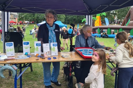 Der Stand des VdK auf der Wiese auf dem M&uuml;Ga Gel&auml;nde. Der Stand ist mit Ulrike Nierhaus (Schriftf&uuml;hrerin) und Ursula Busch (stellvertretende Vorsitzende) besetzt. Informationsmaterial liegt aus. Zwei Kinder spielen und versuchen einen Preis zu gewinnen.