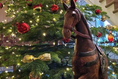 Ein großes, antikes Holzschaukelpferd steht dekorativ vor einem mit großen roten Kugelnd und Lichterkette geschmücktem Weihnachtsbaum.