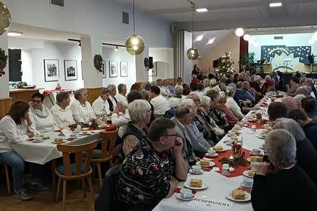 Viele Menschen sitzen in einem Saal an langen, weihnachtlich geschmückten Tischen.