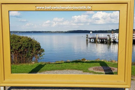 Durch einen großen goldenen Bilderrahmen mit der Aufschrift Bad Zwischenahn Touristik blickt man auf einen See und einen Bootssteg.