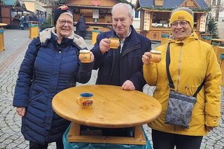 Zwei Frauen und ein Mann stehen an einem Stehtisch aus hellem Holz und prosten mit gelben Glühweintassen in die Kamera.