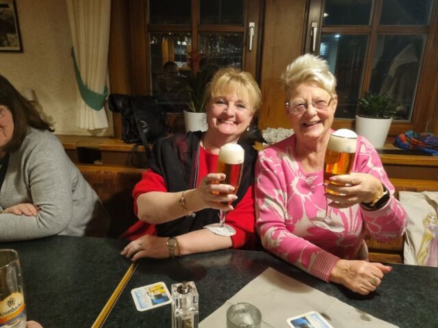 Zwei Frauen sitzen an einem Tisch und prosten lächelnd mit zwei Gläsern Bier in die Kamera.