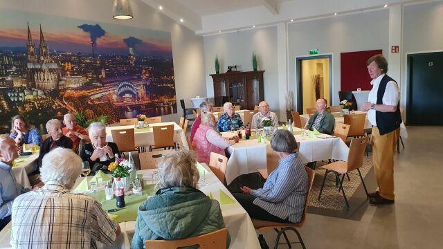 Männer und Frauen sitzen gemeinsam an Tischen in einem Saal. Rechts im Bild OV-Vorsitzender Dr. Martin Happ, der die Gäste begrüßt.