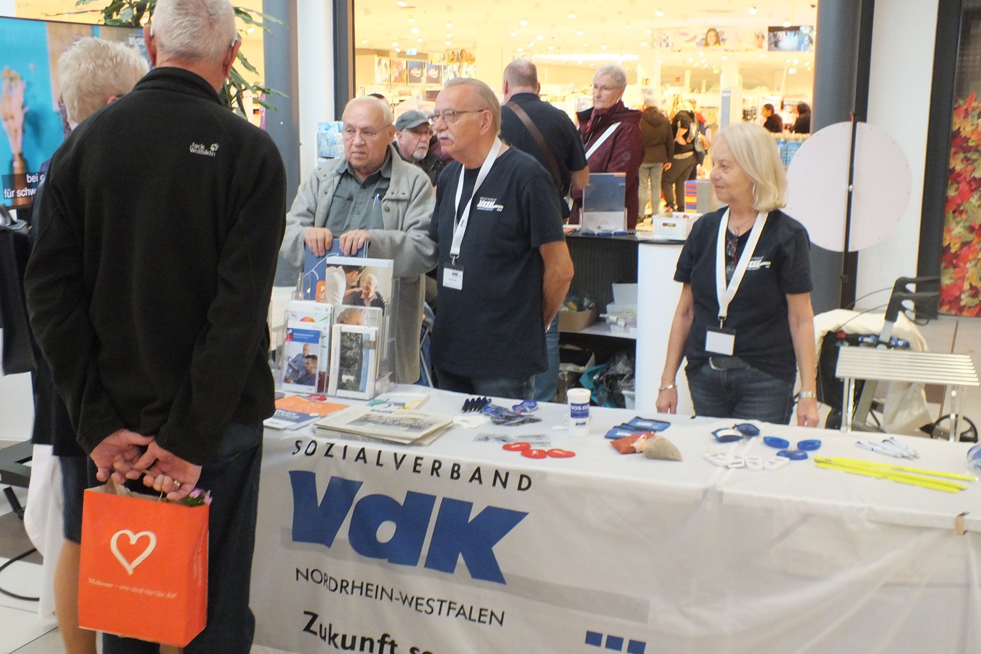 Drei Personen stehen hinter einem VdK Info-Stand und sprechen mit 2 Personen.