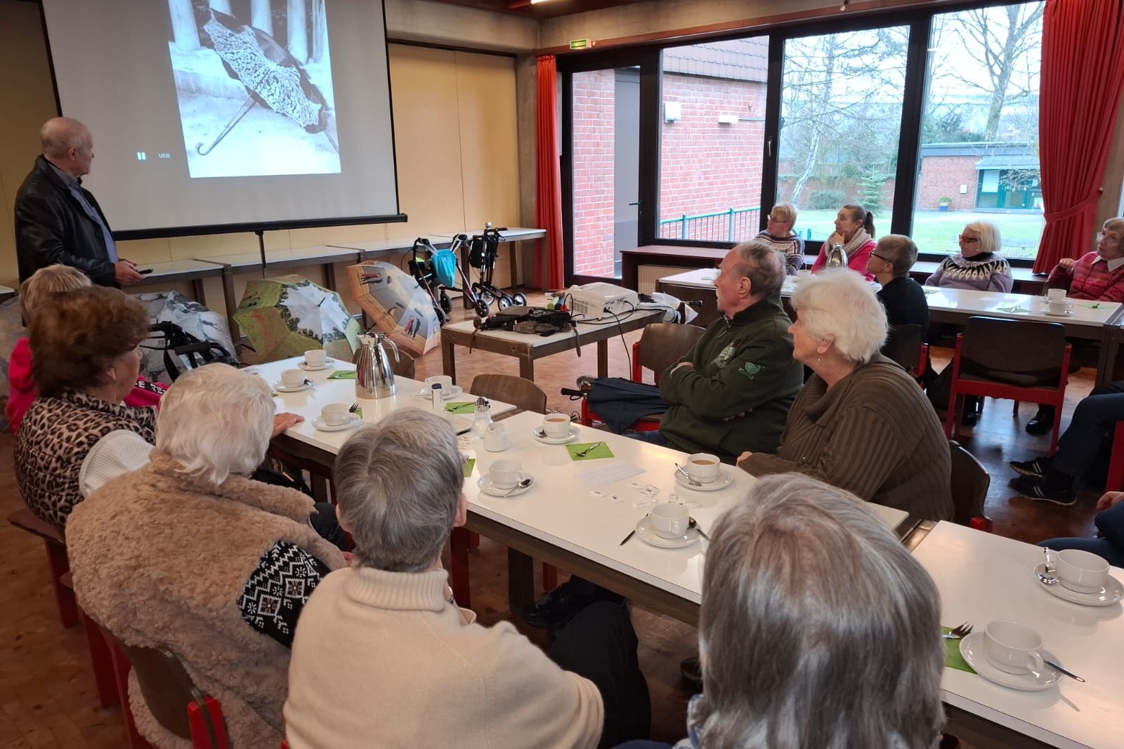 Schirmmacher Willy Schüffler hält einen Vortrag über seinen Beruf und die Geschichte des Schirms. Ein Mann steht vor einer Leinwand und hält einen Vortrag vor einer Gruppe.