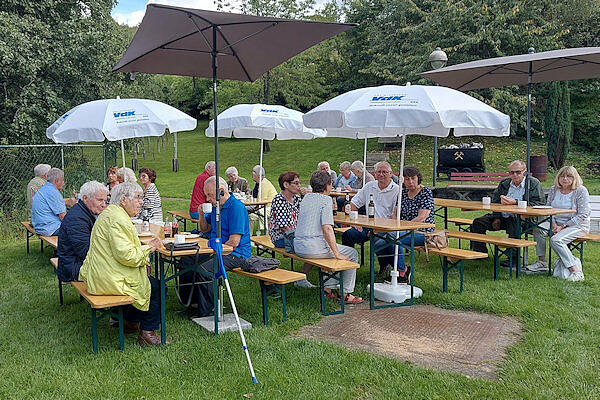 Eine Gruppe Männer und Frauen sitzt an Bierzeltgarnituren, die auf einer großen Wiese aufgebaut sind, unter zwei eckigen und drei weißen Sonnenschirmen mit VdK-Logo. Im HIntergrund steht eine alte Bergwerkslore in der Wiese.