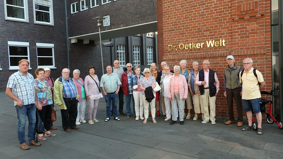 Foto der Reisegruppe vor dem Dr. Oetker Firmensitz unter einem goldenen Schriftzug Dr. Oetker Welt.