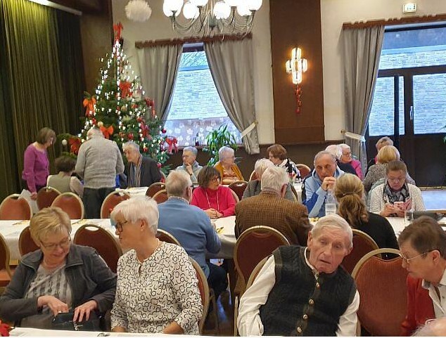 Viele Menschen sitzen an Tischen in einem Saal. Im Hintergrund ein geschmückter Weihnachtsbaum.