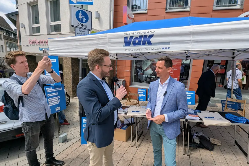 Zwei Männer in Sakko stehen sich vor einem VdK-Pavillon im Gespräch gegenüber. Einer von ihnen spricht in ein Mikrofon. Links im Bild macht ein Dritter mit einem Handy ein Foto.