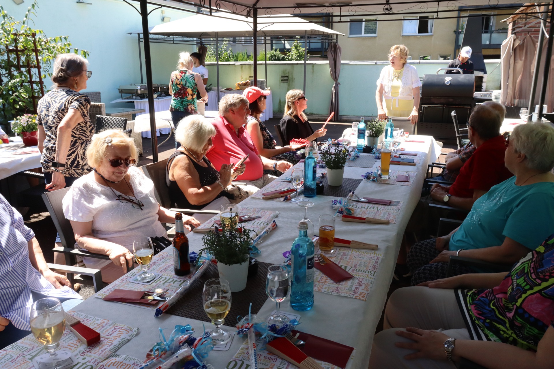Menschen sitzen an langen Tischen auf einer Terrasse bei sch&ouml;nem Wetter. Im Hintergrund steht ein Mann am Grill.