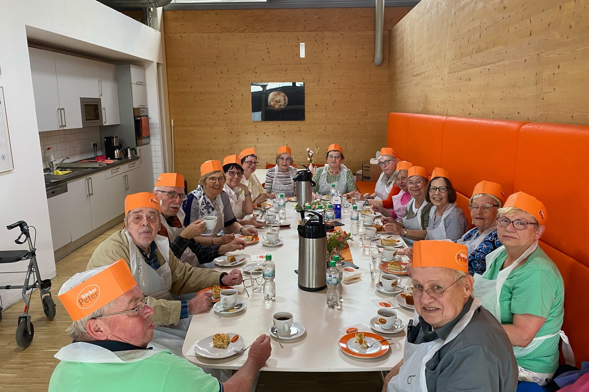 Besuch bei Bäckerei Peter 16 Personen sitzen an einem langen Tisch mit Schürzen und orangen Bäckerhauben bekleidet.
