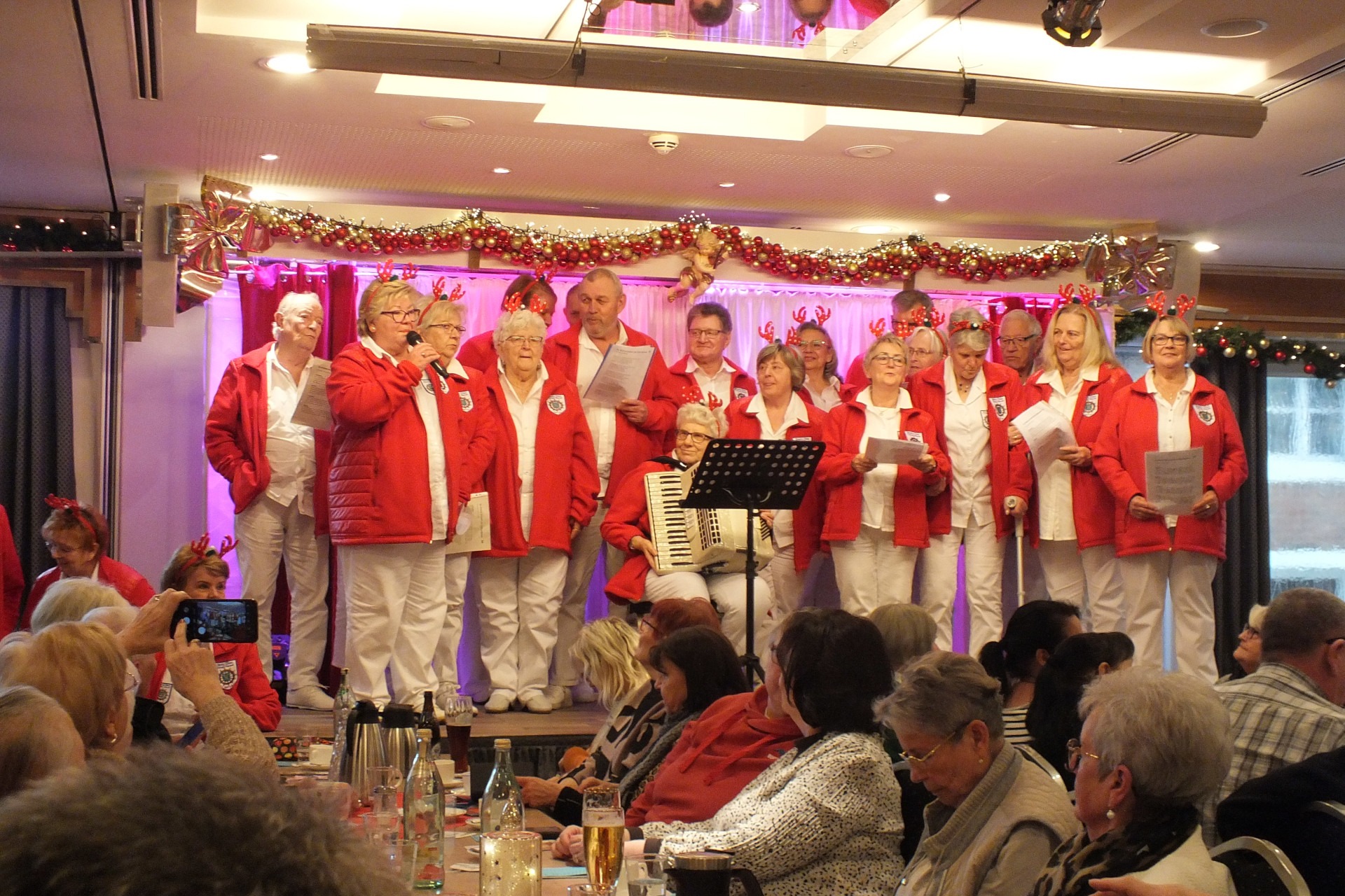 Ein Chor steht auf einer B&uuml;hne in wei&szlig;er Kleidung mit roten Jacken und singen.