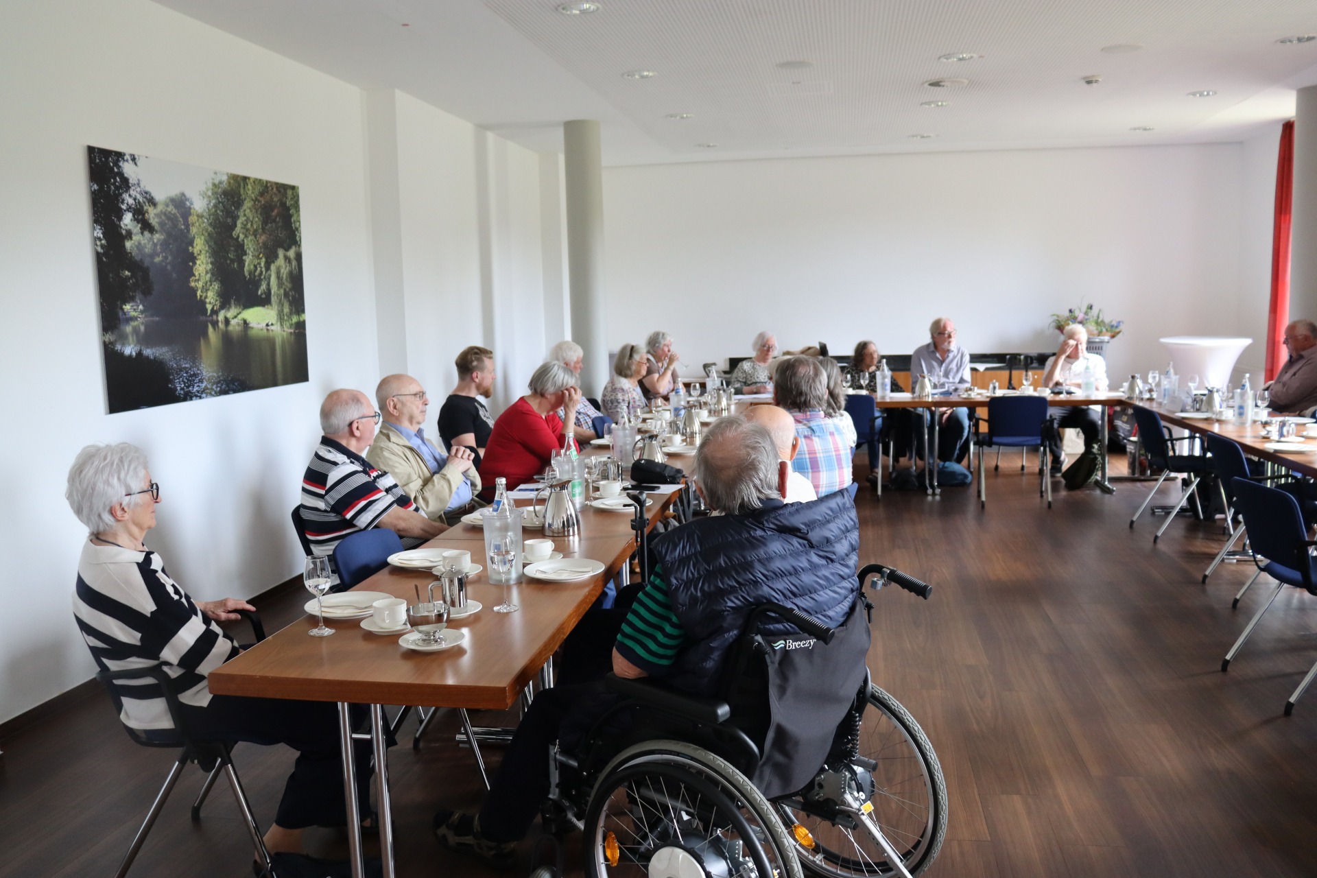 Mehrere Personen sitzen an langen Tischen und h&ouml;ren dem Vorsitzenden zu.