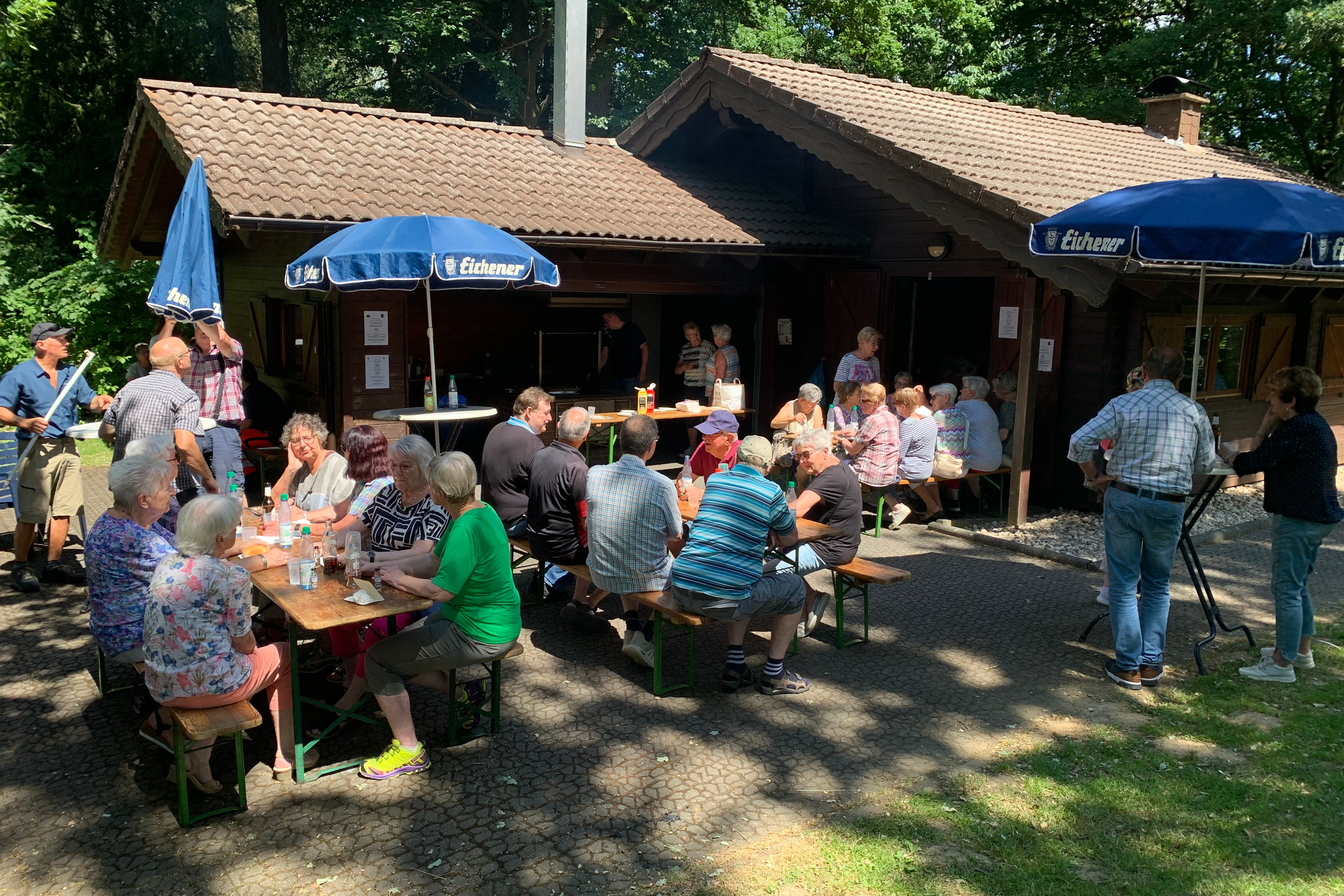 Personen auf B&auml;nken und an Stehtischen vor einer Grillh&uuml;tte im Gr&uuml;nen