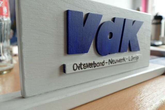 Das Bild zeigt die Buchstaben V d K in blau auf einem grau lackierten Tischschild.