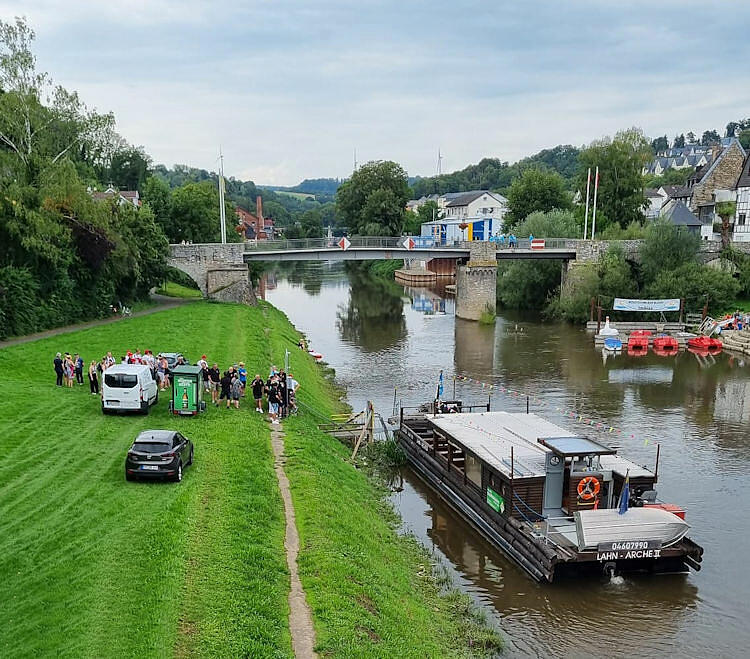 Ein großes Floß liegt vertäut am Ufer der Lahn. Daneben warten ein Transporter und ein PKW und einige Menschen.