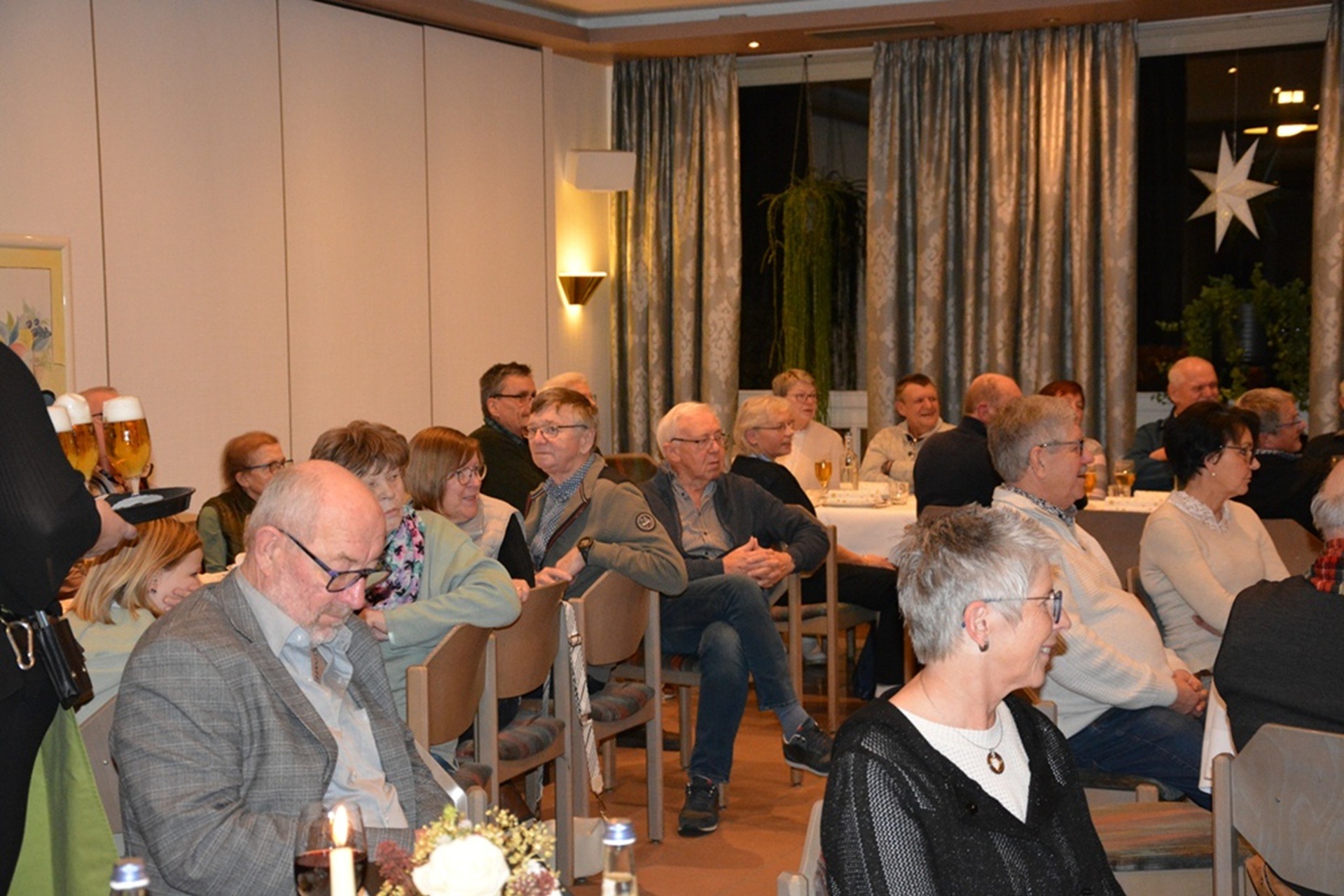 Viele Menschen sitzen gemeinsam in einem weihnachtlich dekorierten Saal an Tischen