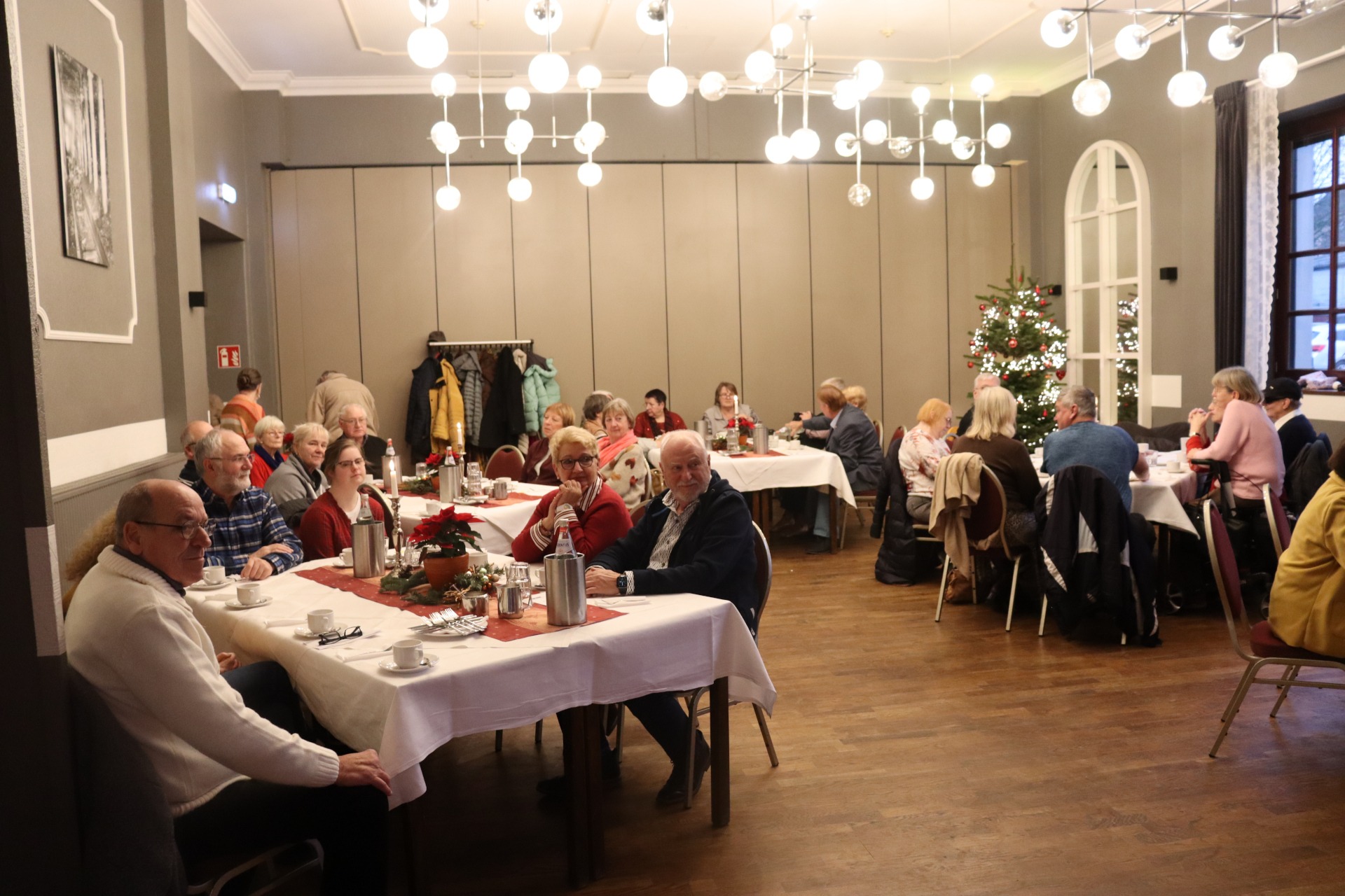 Mehrere Mensche sitzen an festlich gedeckten Tischen. im Hintergrund steht ein geschm&uuml;ckter Tannenbaum.