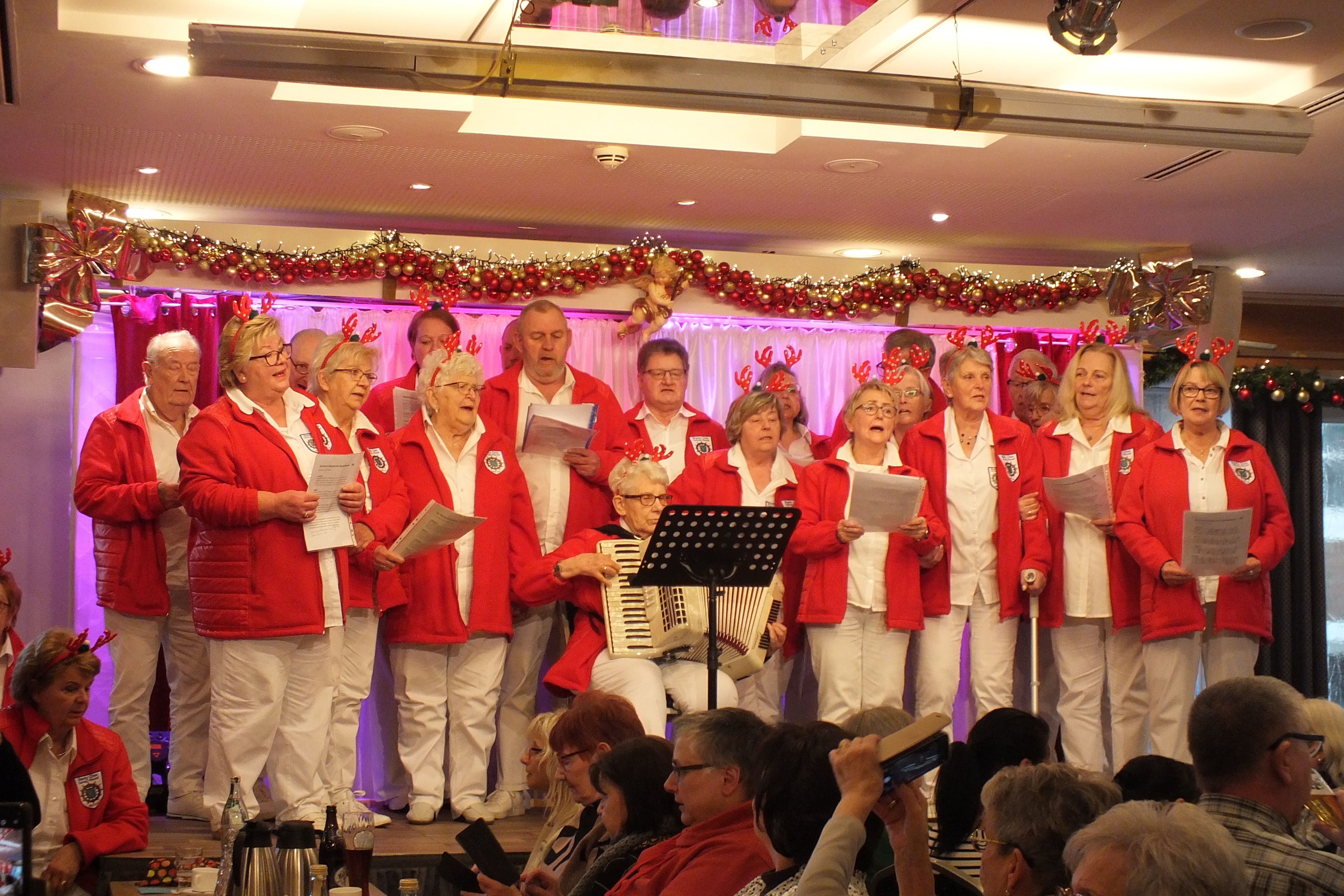 Ein Chor steht auf einer B&uuml;hne in wei&szlig;er Kleidung mit roten Jacken und singen.