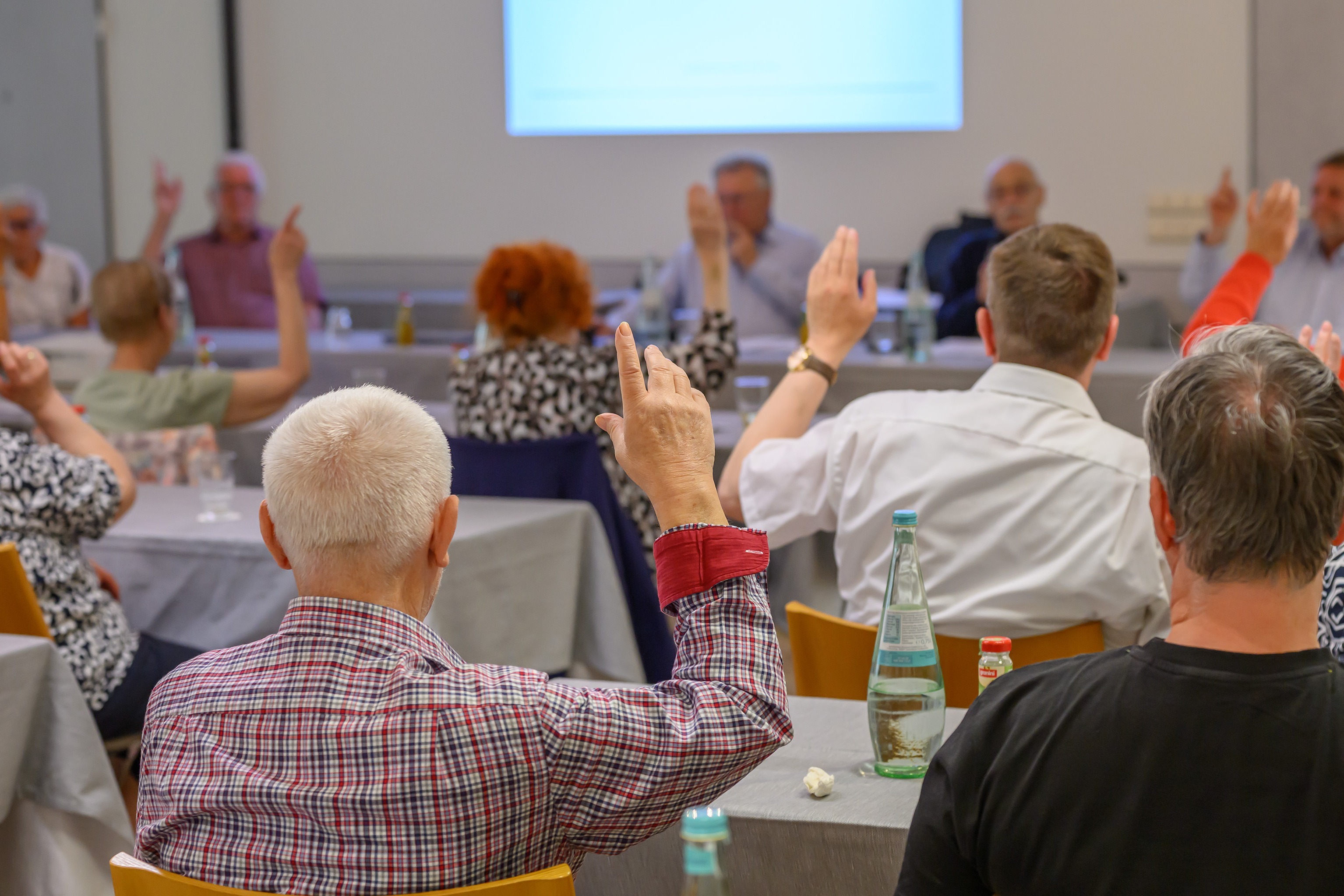Menschen sitzen an Tischen in einem Saal und heben die Hand zur Abstimmung.