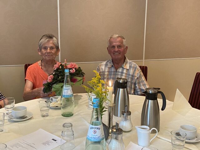 ein Mann und eine Frau sitzen an einem runden, gedeckten Kaffeetisch. Die Frau hat einen Blumenstrauß vor sich stehen.