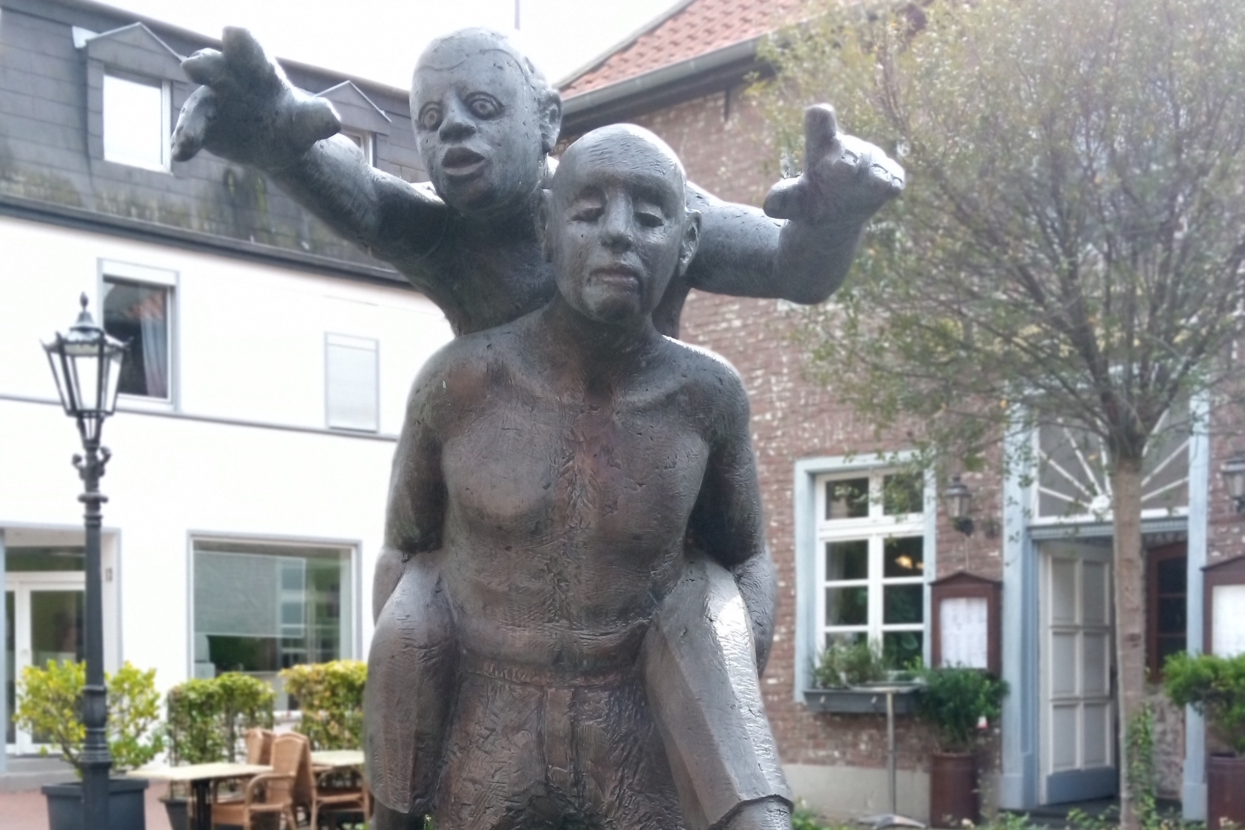 Skulptur "Der Blinde und der Lahme" Die Skulptur in Viersen-Dülken zeigt einen blinden Mann, der einen Mann auf dem Rücken trägt. Der Mann auf dem Rücken zeigt nach vorne und sagt dem Blinden den Weg.