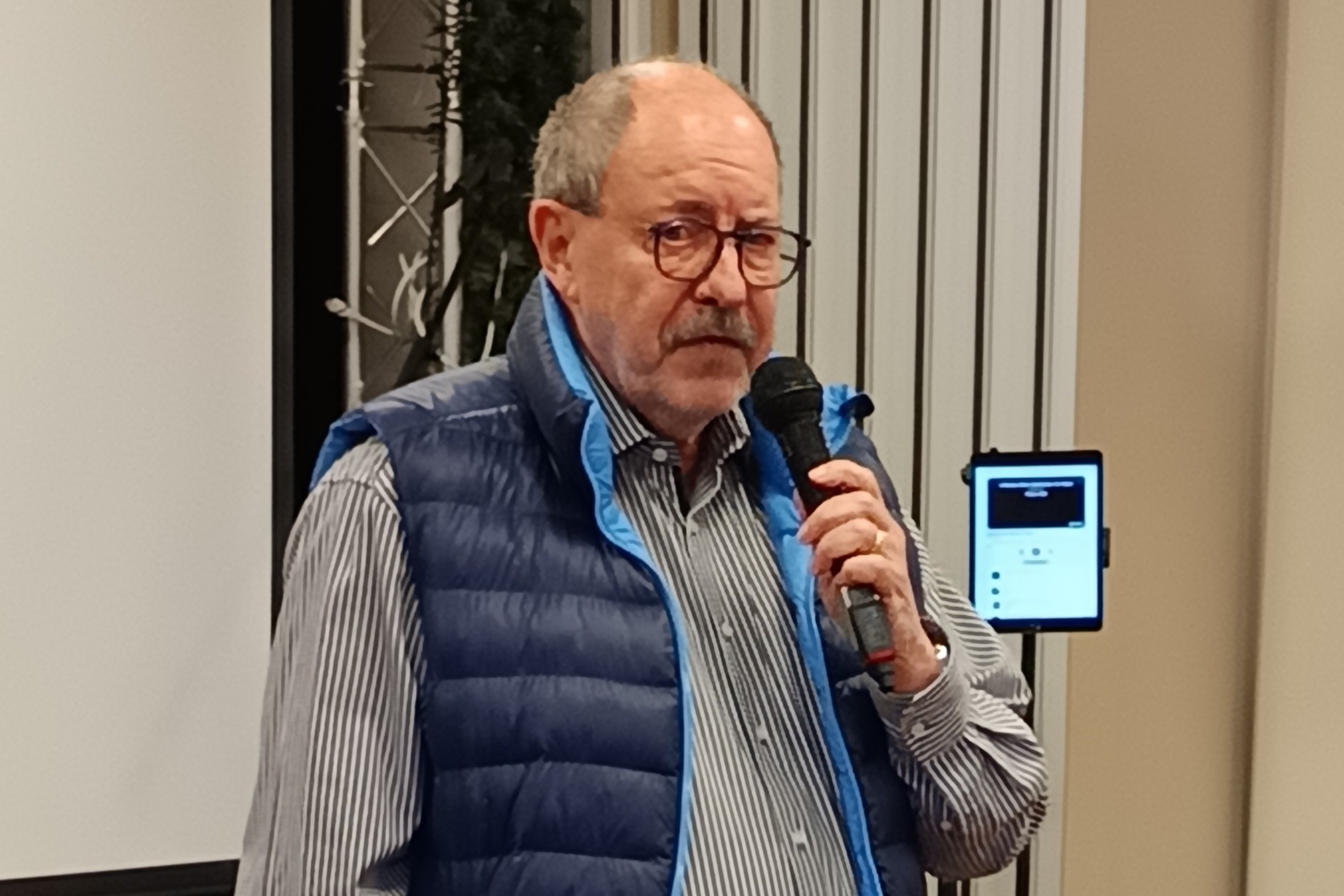 Auf dem Bild sieht man den Vorsitzenden des VdK OV Bilk, Man sieht einen &auml;lteren Herren mit Brille und blauer Weste mit einem Mikrofon in der Hand.