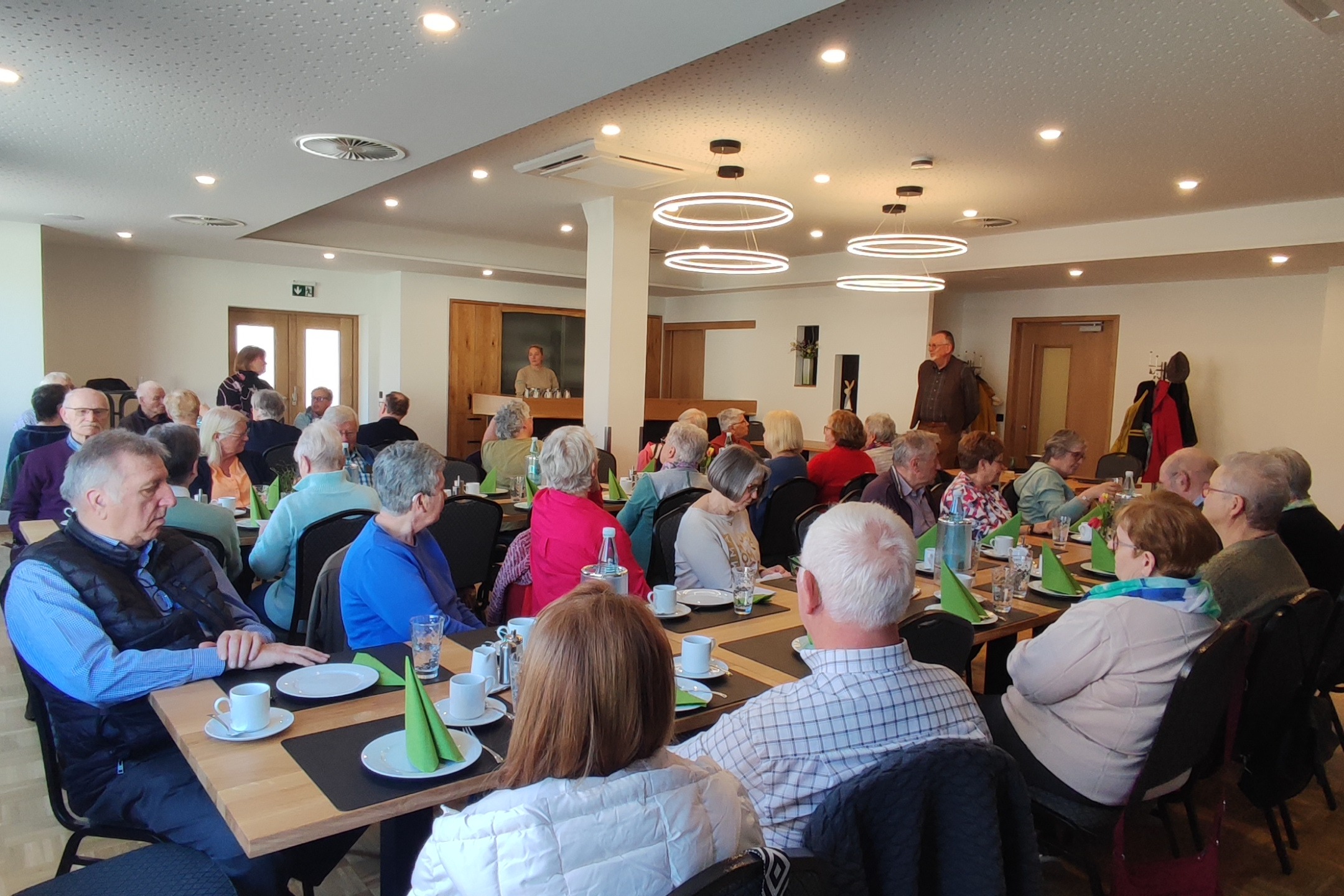 Viele Menschen sitzen an gedeckten Tischen in einem Raum. An der rechten Seite steht ein Mann und spricht zu Publikum.