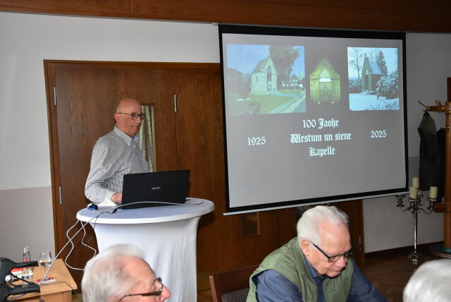 Ein Mann steht an einem Stehtisch neben einer Leinwand, auf der Beamer-Fotos der Westhumer Kapelle zu sehen sind.