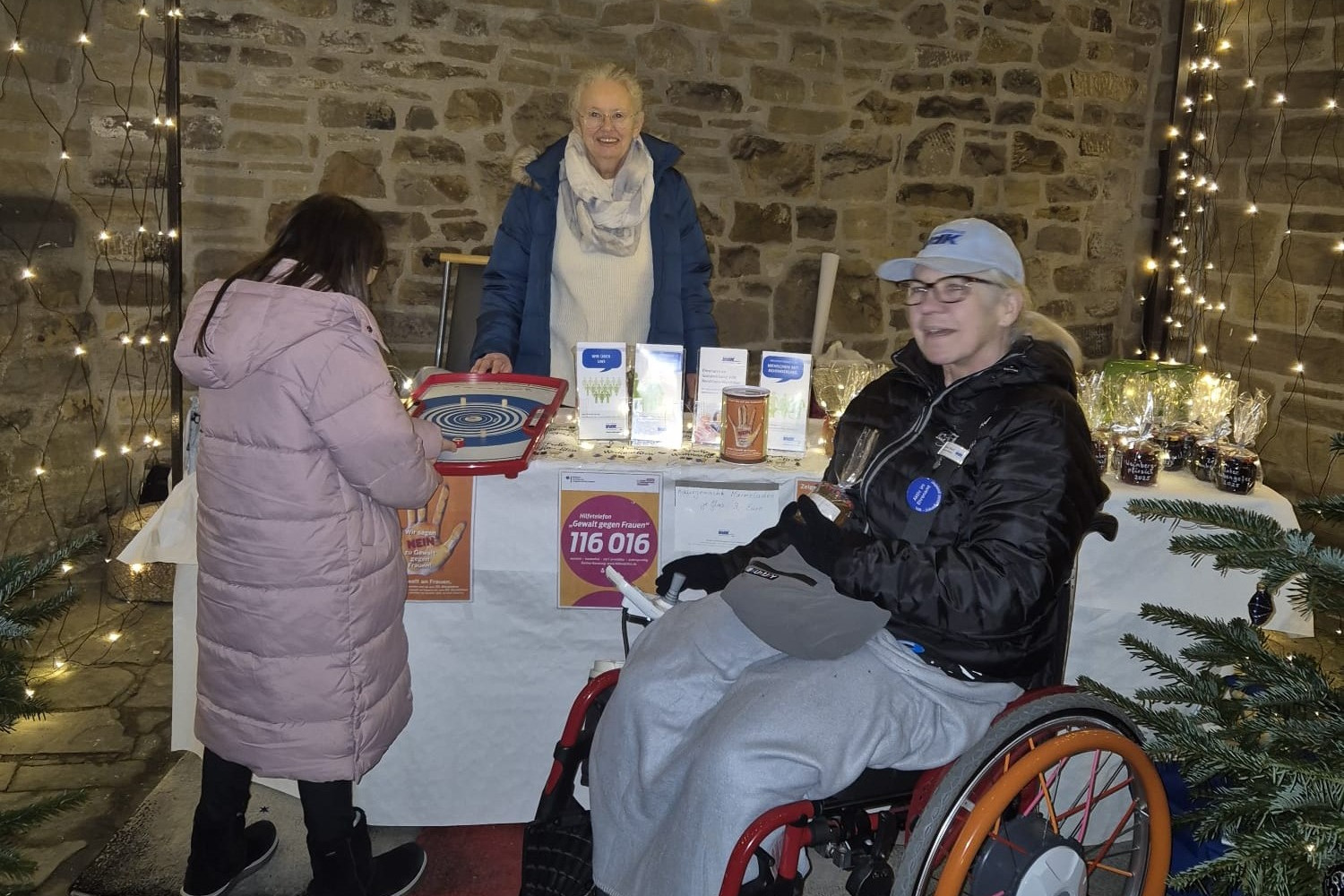 Informationsstand des VdK Kreisverbandes M&uuml;lheim an der Ruhr auf dem Adventsmarkt 2025 in der M&uuml;lheimer Altstadt. Man sieht wie die Frauenbeauftragte Christel Braun und die stellvertretende Vorsitzende Ursula Busch Informationsmaterial anbieten und ein Kind am angebotenen Tischspiel versucht einen Preis zu gewinnen.