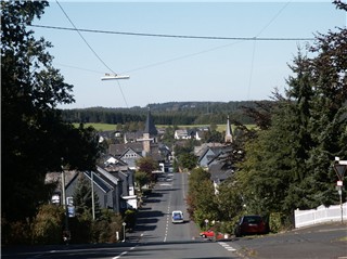 Blick auf die Ortschaft Wilnsdorf mit einer breiten, von Fichten gesäumten Straße, die hügelabwärts führt, den Dächern des Ortes und zwei Kirchtürmen.