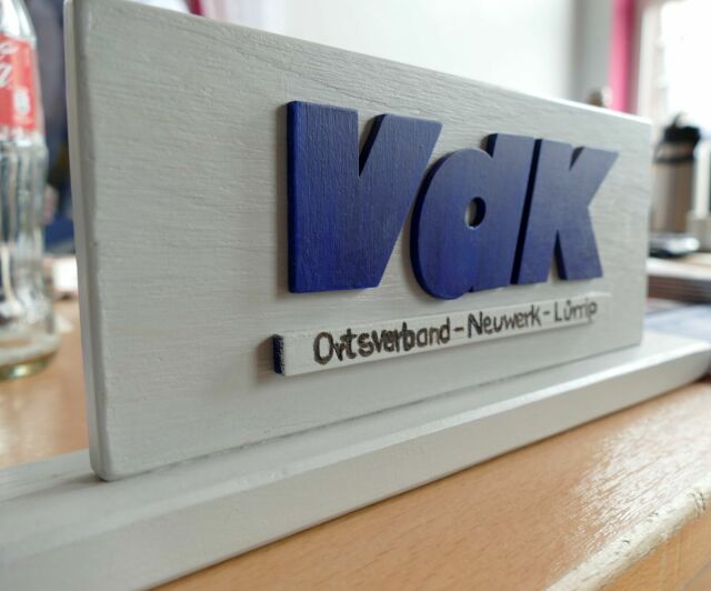 Das Bild zeigt ein graues Tischschild mit dem Schriftzug VdK in blauer Farbe und Ortsverband Neuwerk Lürrip darunter geschrieben in schwarzer Farbe.